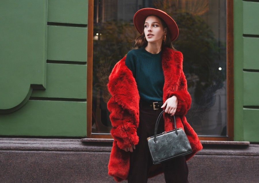 Outdoor fashion tips portrait of young beautiful confident lady wearing trendy orange faux fur coat, hat, green sweater, holding stylish snakeskin textured handbag, posing in street of city.
